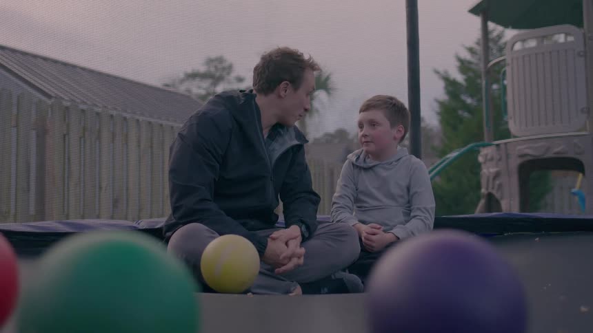 Young man and boy converse while sitting on a trampoline in a play area at sunset