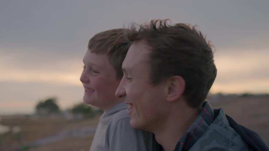 A smiling man and boy look out into the distance together during a peaceful sunset