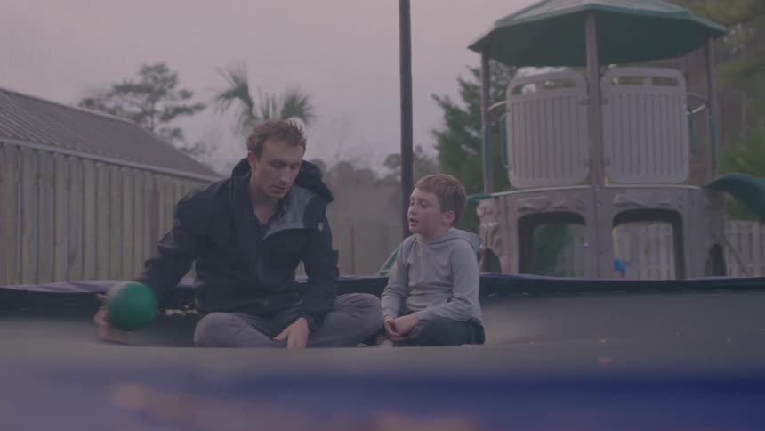 A man and a boy sit on a trampoline having a heartfelt conversation in a backyard at dusk