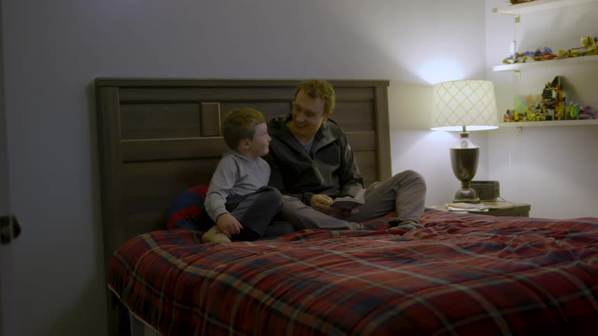 A father and son sit on a bed in a cozy bedroom sharing a quiet bonding moment