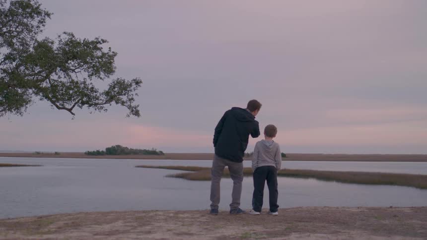 A father and son stand on a lakeshore quietly admiring the peaceful sunset view