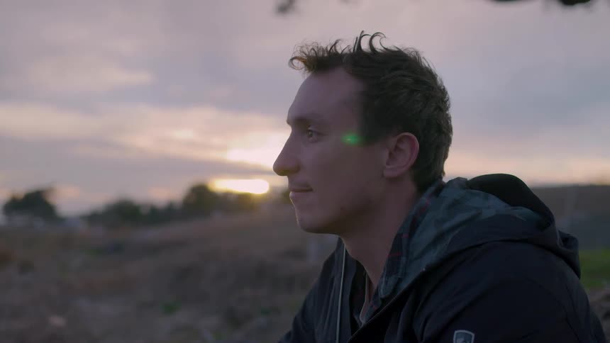 A man sits outdoors at dusk looking thoughtfully into the distance