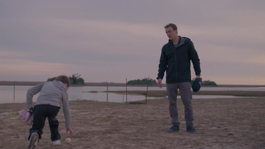 A father and son play catch with baseball gloves near a lakeside path in the evening light