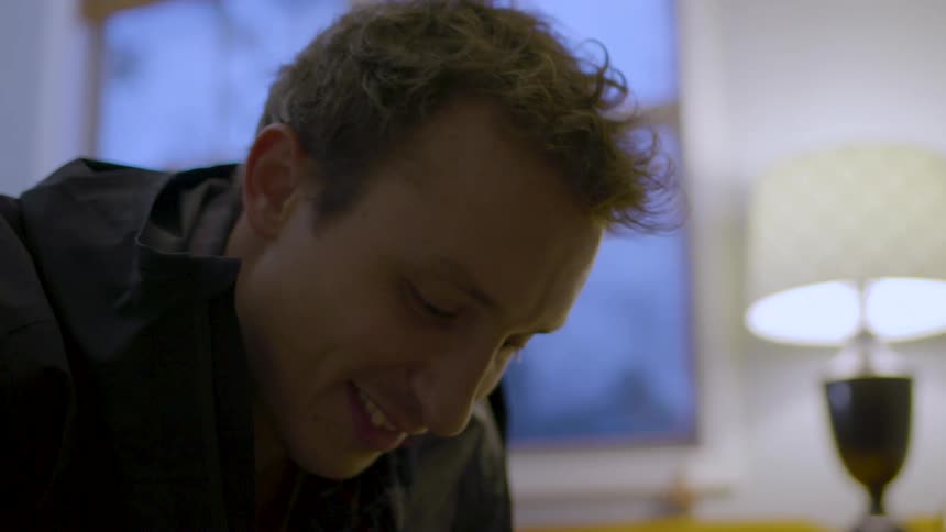 The face of a man smiling while looking at something below him in an indoor setting with a small light on behind him