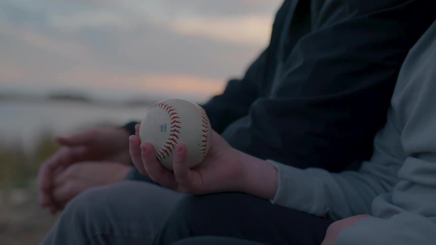 Close up of a boy holding a baseball while sitting at the riverbank with an adult at sunset
