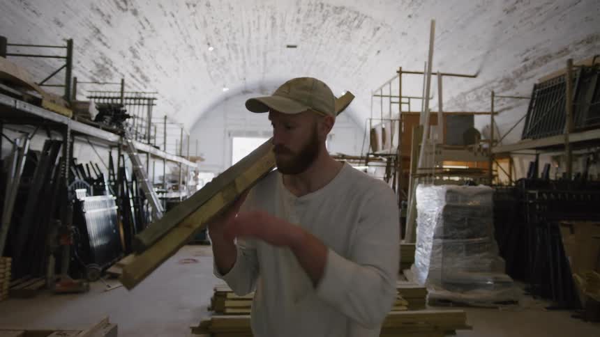 A man in work clothes carries wood planks on his shoulder inside a large industrial workshop