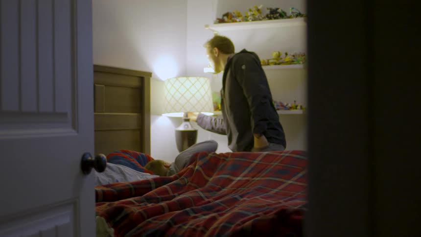 A father kisses his son goodnight in a warmly lit bedroom before turning off the lamp