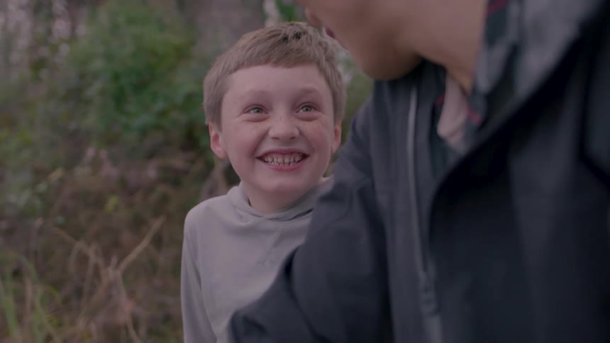 A young kid laughs while talking to a man wearing a black jacket in the woods at midday