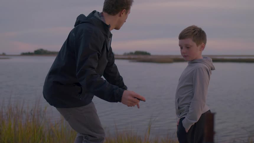 A man standing next to a little boy is hitting a stone on the surface of a lake making it bounce