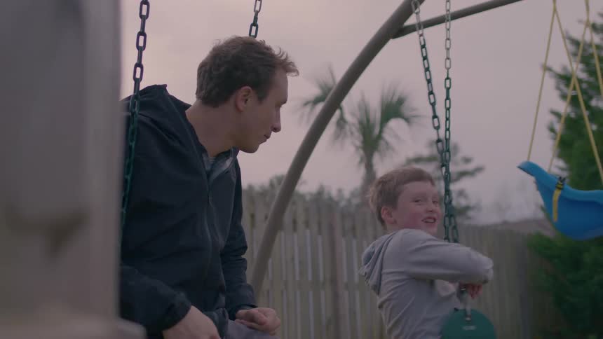 Young man and a boy play and converse on a swing in a playground outdoors at sunset
