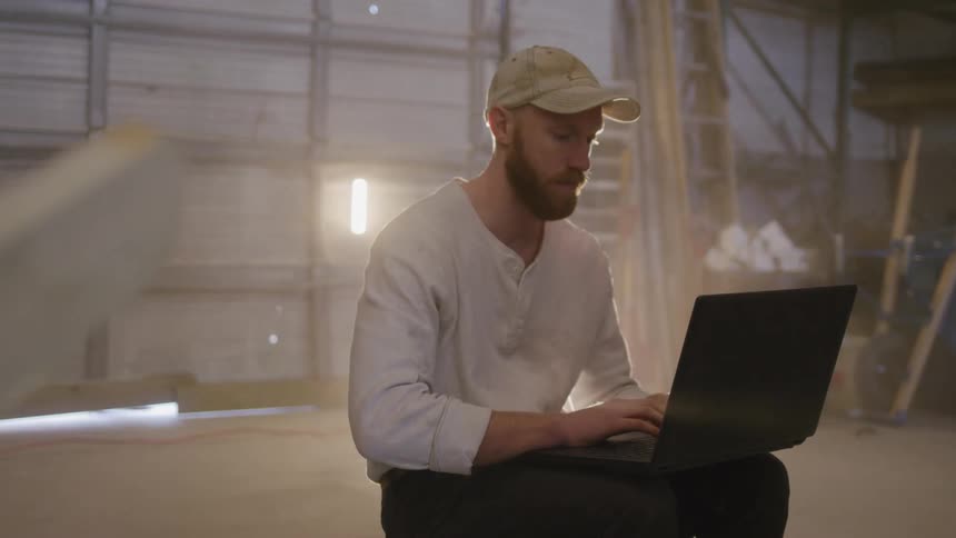 A man in casual work clothes uses a laptop inside a dusty workshop