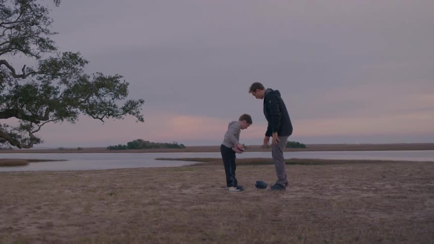 A man and a boy outdoors with the man placing a baseball into the boys hand who is wearing a baseball glove