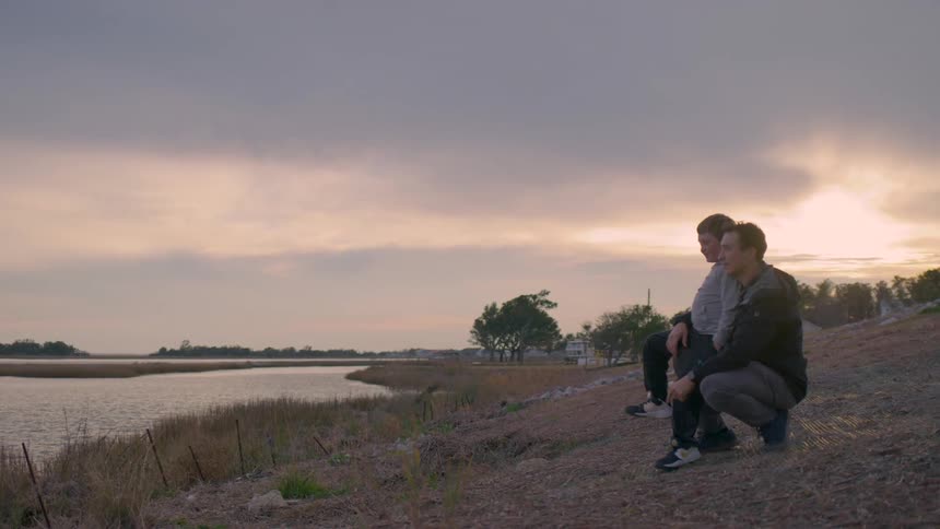 A father and son sit quietly by a riverbank at sunset