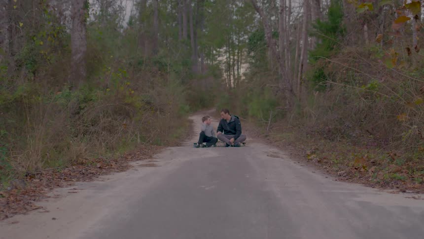 A man and a child sit in the middle of a road surrounded by forest at midday