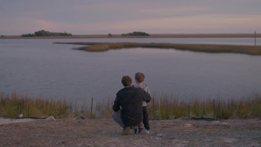 A father and son kneel by a calm lakeside gazing at the water during dusk