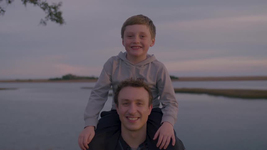 The boy is sitting on his fathers shoulders by the shore