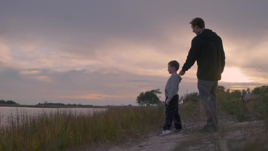 Boy stands and laughs with a young man at the river bank at sunset while playing stone skipping