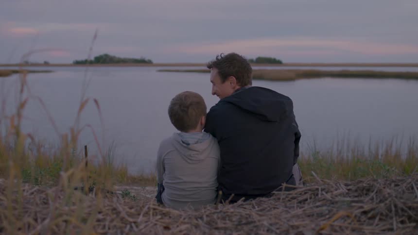 A man and child sit close together by a lakeside sharing a quiet moment at sunset