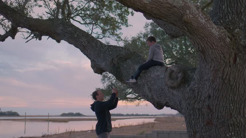 Father photographs his son sitting on a large tree by the water at sunset
