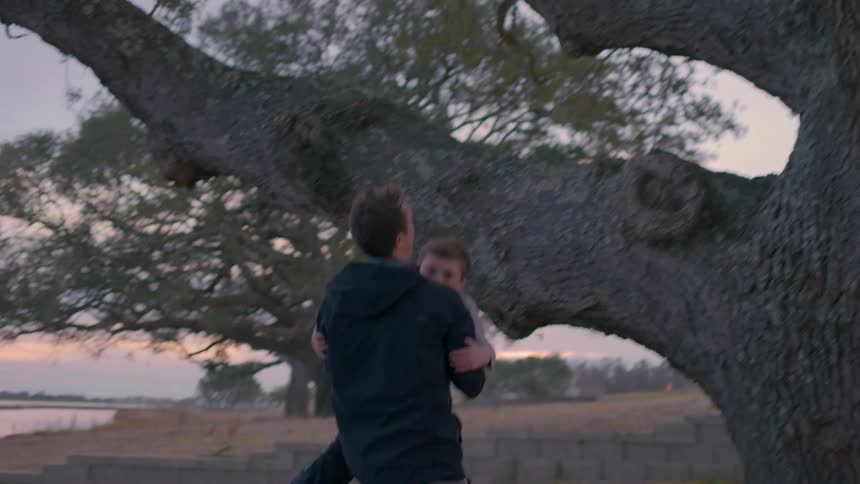 Young man catches a boy that jumps down from a tree branch next to the river at sunset
