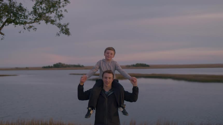 A man gives a child a shoulder ride as they pose for the camera near a calm body of water at sunset