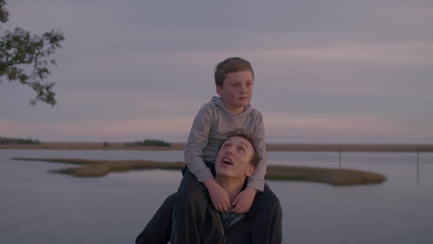 A father walks near water at sunset with his son riding on his shoulders