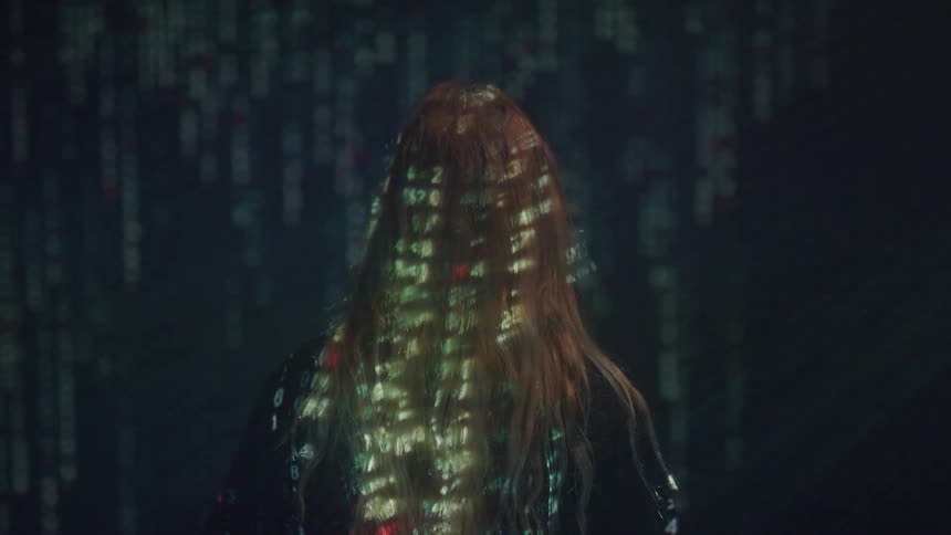 A person stands in front of a projector screen displaying matrixlike numbers with the projection casting onto their body as they use a cloth to cover their head