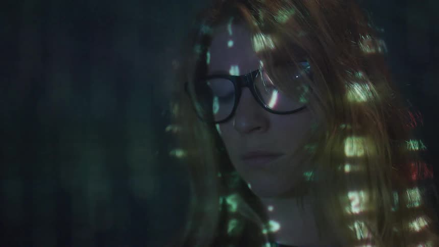 A blonde woman wearing glasses sits in a dark room illuminated by the glow of numbers from a screen reflecting across her face