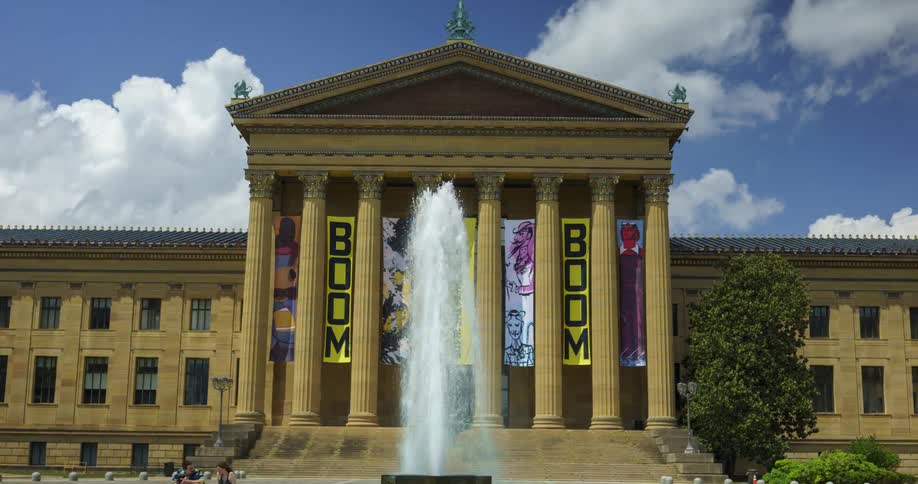 A hyperlapse of The Philadelphia Art Museum with a fountain in front of it as people walk in and out of the building at midday
