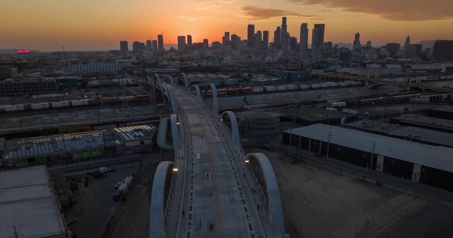 A city skyline view at sunrise shows the Sixth Street Bridge Viaduct in Los Angeles with a series of cars passing by