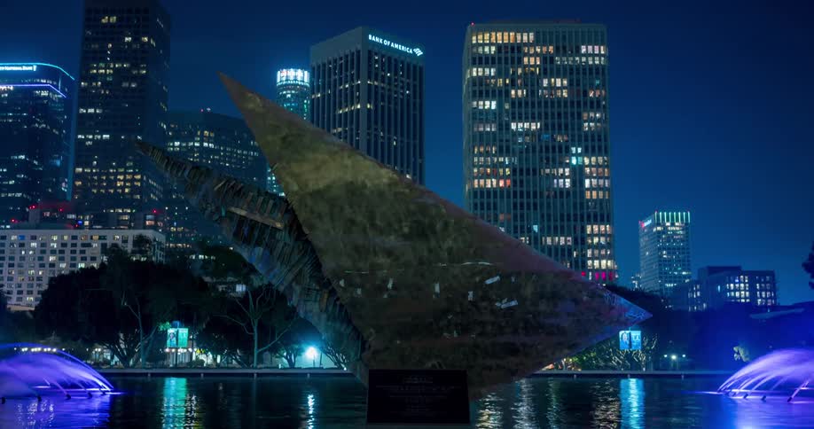 A triangular sculpture glows with purple and blue lights beside a fountain framed by tall modern buildings at night in Los Angeles