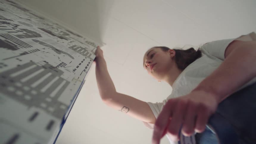 A young woman applies paper tape to secure wallpaper glued to a wall at midday