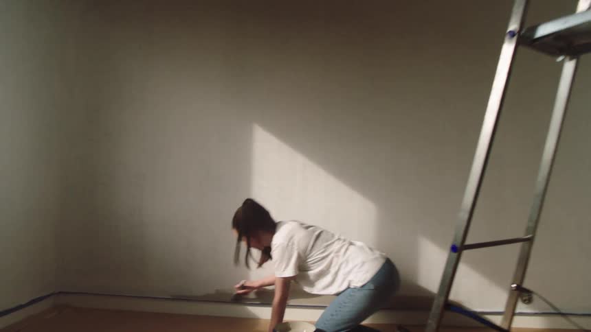 Young woman kneels while painting the bottom area of a room wall during daytime