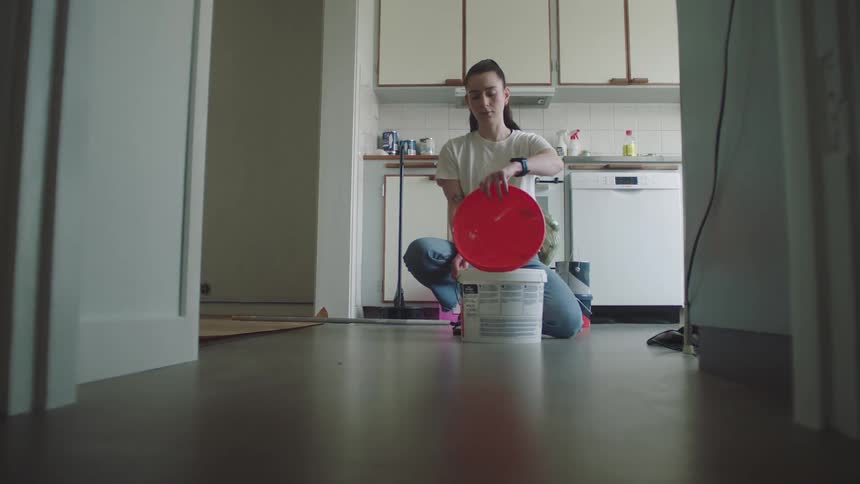 Woman kneels in kitchen opening red bucket to pour paint