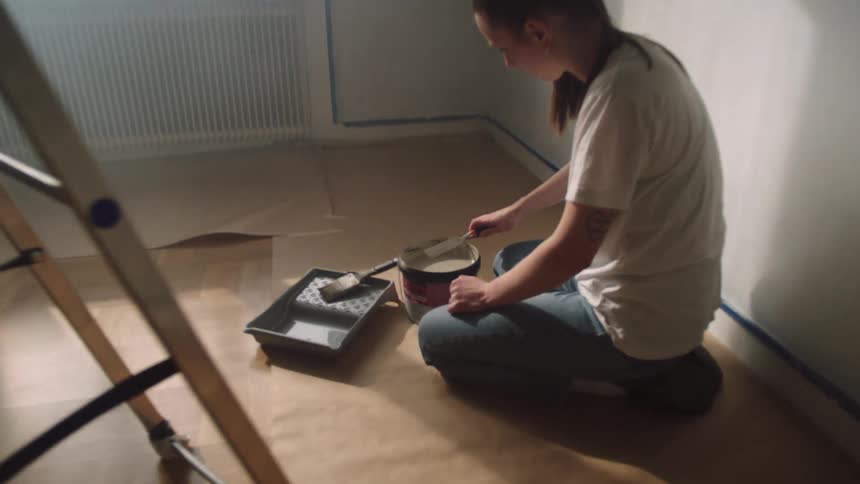 Female home painter kneels in a room while mixing paint in a container