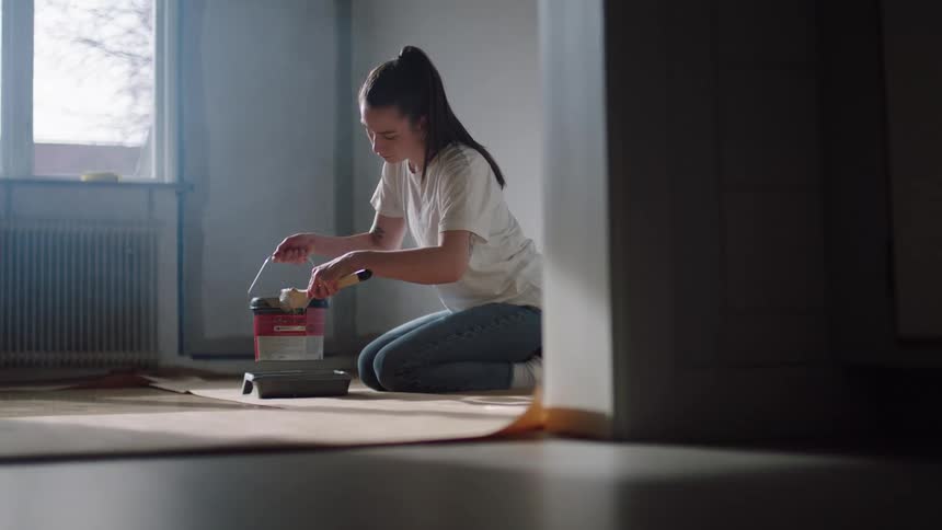Woman kneels on the floor dipping a paint roller into a tray inside a sunlit room