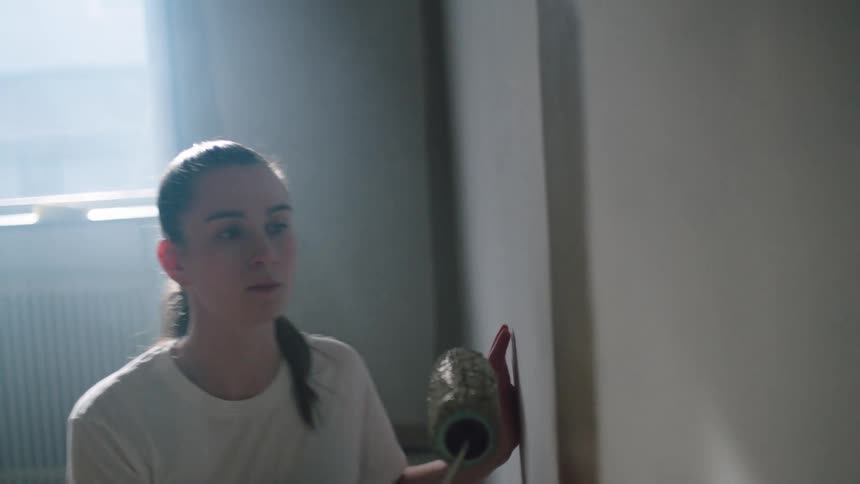 A woman crouching on the floor is putting paint on the brush roller and painting a white wall