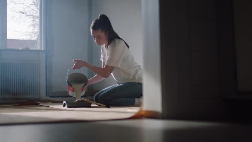 Young woman carefully pours paint into a paint tray while kneeling in a room