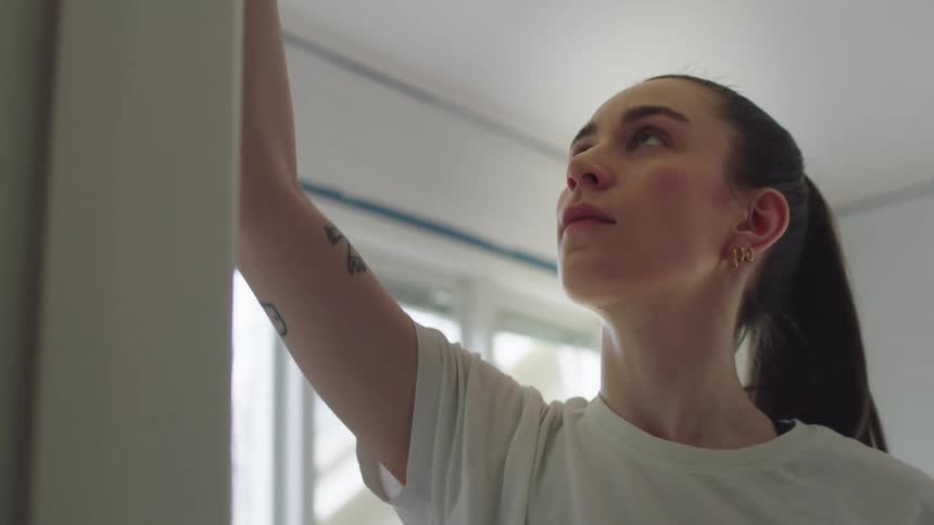 A young white woman uses a brush to apply paint to an interior building wall at midday