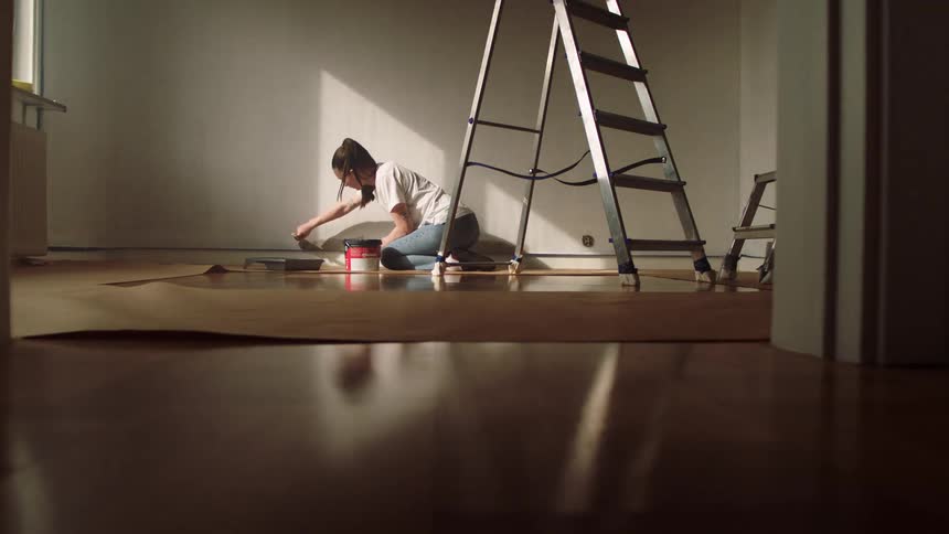 A woman paints a wall while kneeling on the floor in a sunlit unfurnished room