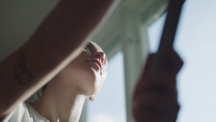 A young woman inside a room rolls paint up and down a wall at midday
