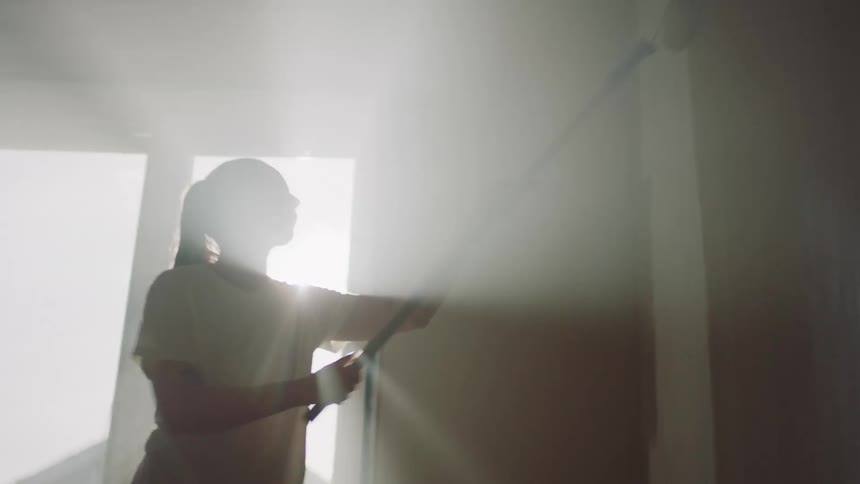Woman paint rolling an interior wall as the sunlight flares behind her