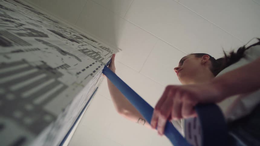 Young woman applies blue tape vertically on the edge of a room wall