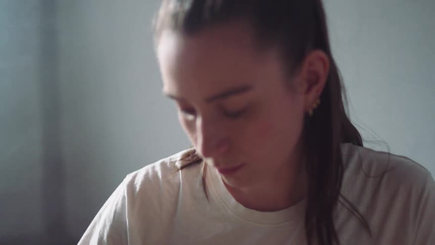 A woman wearing a white shirt with long hair tied back sits in a softly lit room