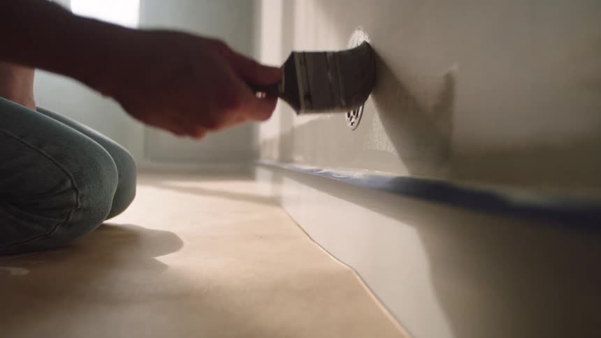 A person kneels on the floor while painting a wall with a brush near a drain cover
