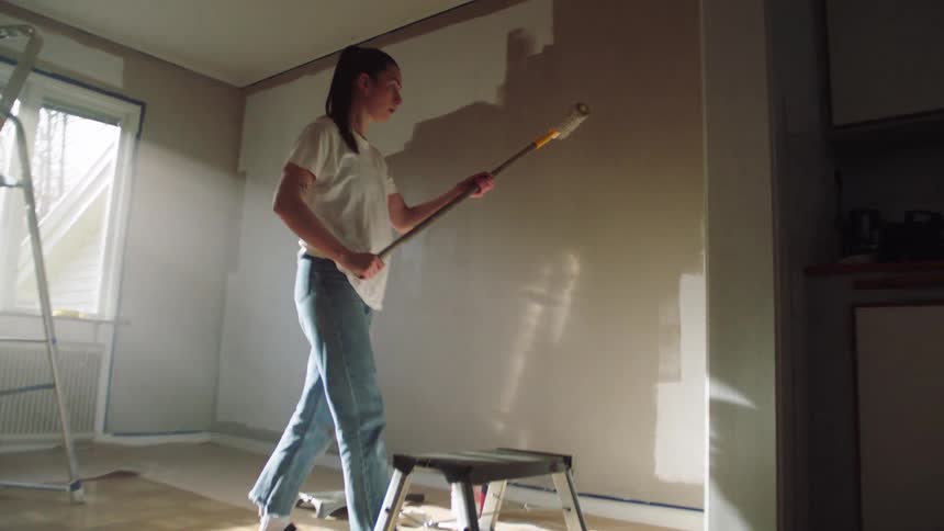 In a sunlit room a woman uses a roller on an extension pole to paint a wall white