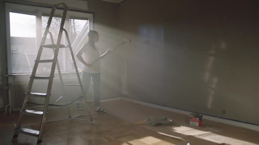 A young woman wearing a white tshirt and jeans uses a roller to paint the wall of a room at midday with a ladder positioned behind her