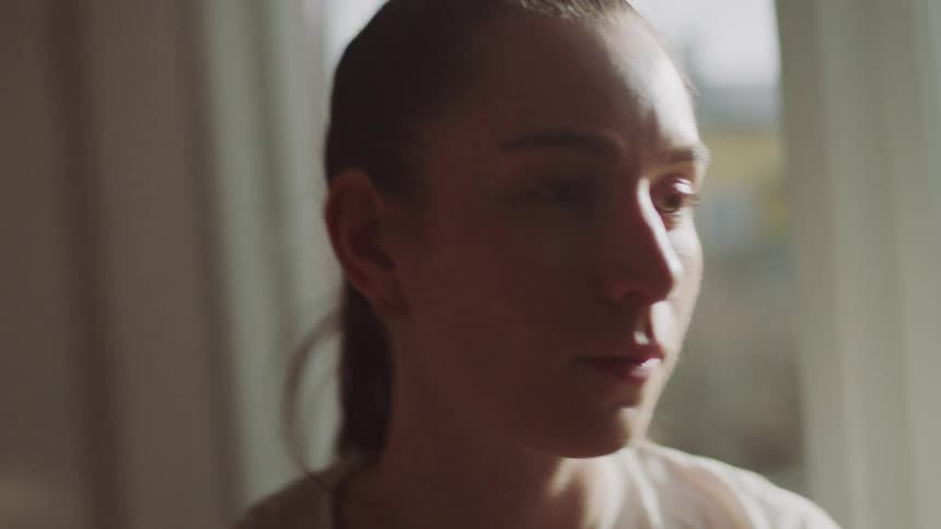 The face of a young woman with her hair tied back and she is looking at the wall that is in front of her next to a window and her face is being illuminated by natural light