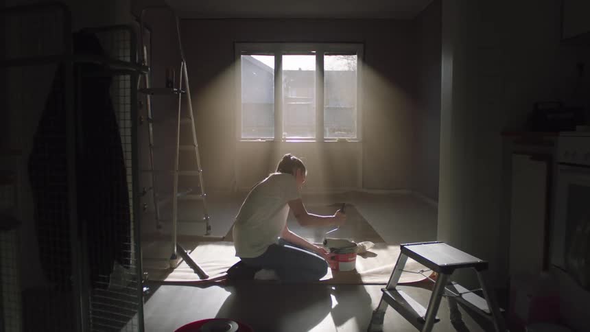 A person kneels on the floor in soft sunlight preparing to paint a room
