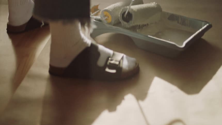 A woman in sandals and socks dips a paint roller into a tray of white paint on a concrete floor preparing to paint a brightly lit room